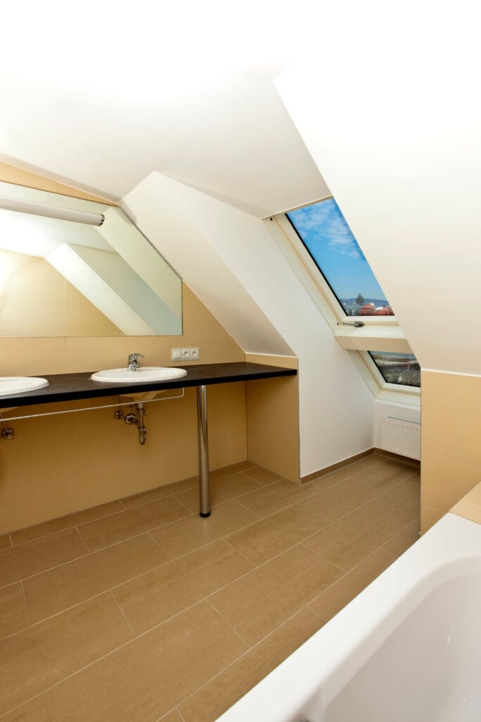 一间窄窄的米色阁楼浴室，带有一个工业风格的梳妆台，内有两个传统的水槽。右边是一个带天窗的小凹室。