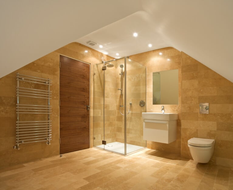 金色瓦片倾斜屋顶下的小浴室。淋浴间、水槽和便盆排列在远处的墙上，与门和毛巾架相邻。