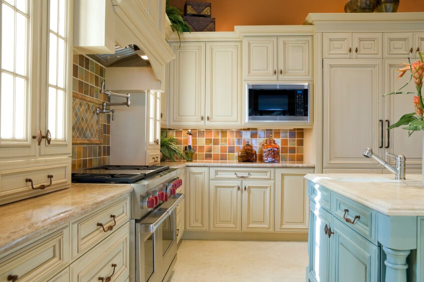 一个乡村厨房，带有浅蓝色的岛台，后挡板上贴着五颜六色的瓷砖。与这个系列中的许多其他厨房一样，炉灶上方的一个协调的中心装饰物为后挡板的大部分增加了视觉上的乐趣。