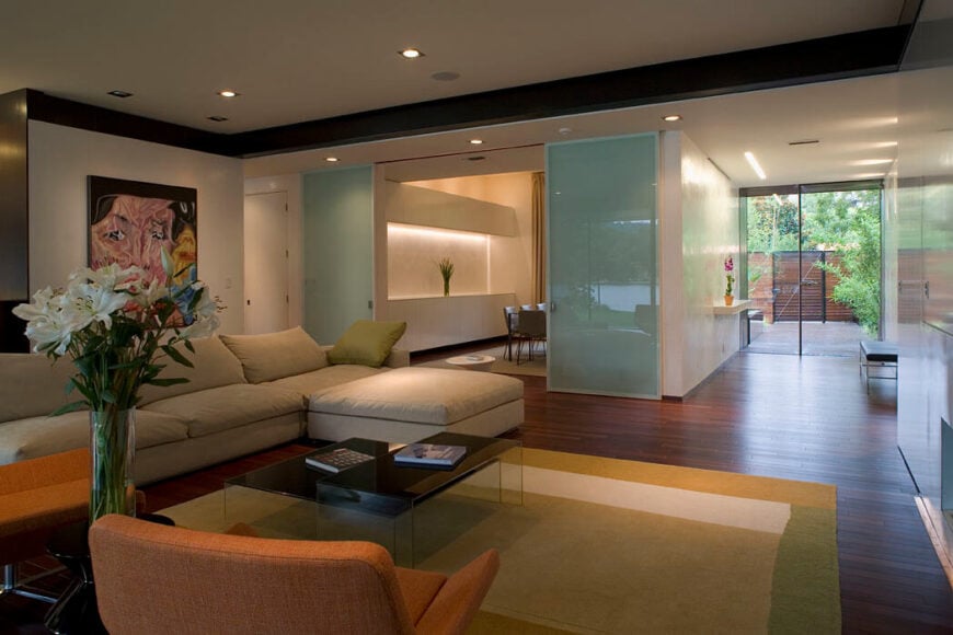 客厅铺着丰富的硬木地板，贯穿整个住宅，覆盖着绿色和白色的地毯。奶油色组合沙发搭配着两把亚麻橙色的现代扶手椅。滑动光滑的蓝色玻璃门打开，露出餐厅。