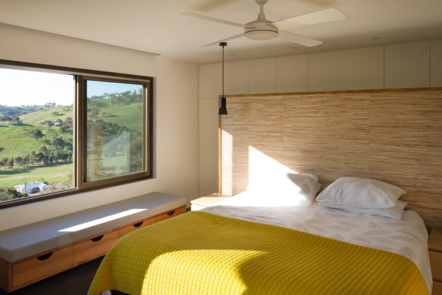 设计简单的卧室有一个有质感的床头板和黄色的被子。大窗户下面放着一张储物凳。