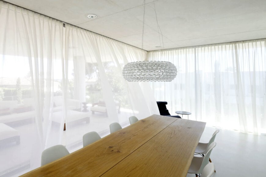 较低的餐厅设有一个大的天然木桌，周围是定制的白色椅子，在一个完全向室外开放的空间里。透明的窗帘环绕着房间。