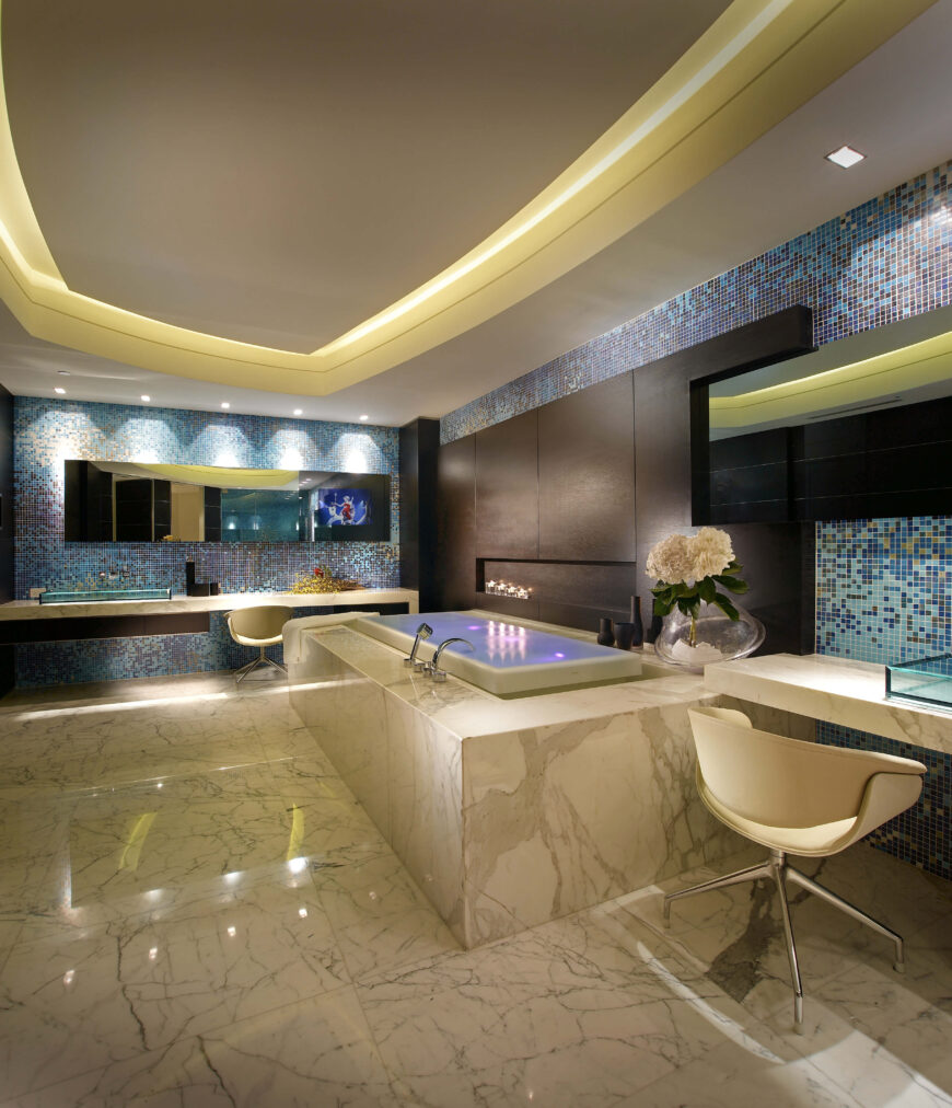 宽敞的主浴室里有一个巨大的浴缸、化妆台和蓝色和棕色色调的瓷砖墙壁。托盘天花板是围绕四周照明的。
