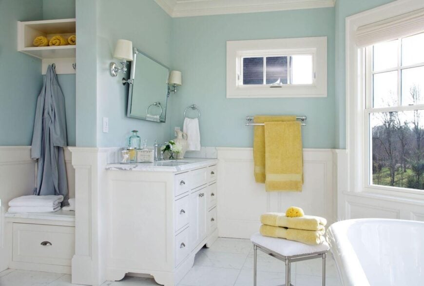 一个白色和知更鸟蛋蓝色的浴室与阳光黄色的口音。有一个带收纳钩和一个拉出抽屉的小长凳可以作为额外的毛巾收纳处。壁挂式镜子可以调节到完美的角度。