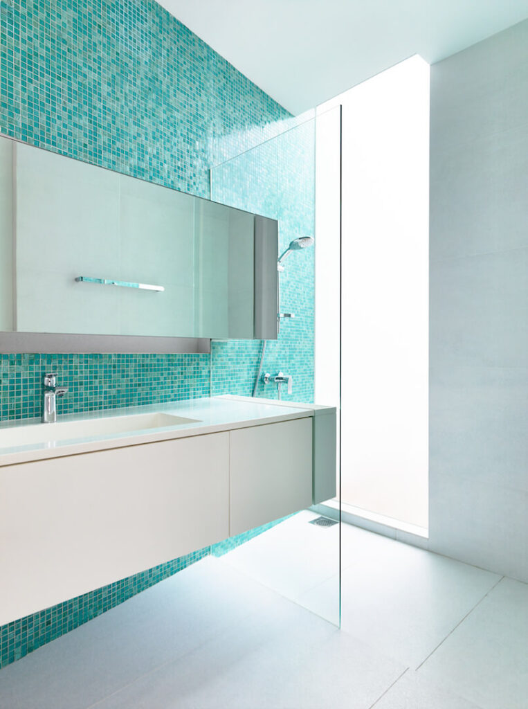 一个更现代的浴室，没有以前的浴室的自然特征。漂浮的梳妆台后面的蓝色落地玻璃瓷砖与原始的地板、梳妆台和其他三面墙形成了鲜明的对比。淋浴间的落地窗可以让人从隐私屏幕后面欣赏露台。