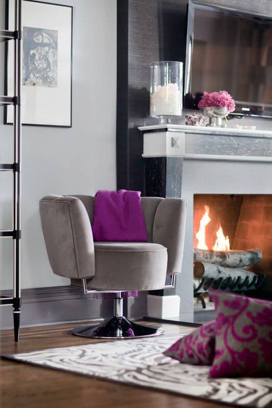 客厅的燃木壁炉和旁边的现代米色麂皮椅子的特写。圆形椅子的靠背上挂着一条漂亮大胆的浆果紫色流苏羊绒毯子。壁炉周围美丽的黑色皂石增添了一种优雅的气氛。