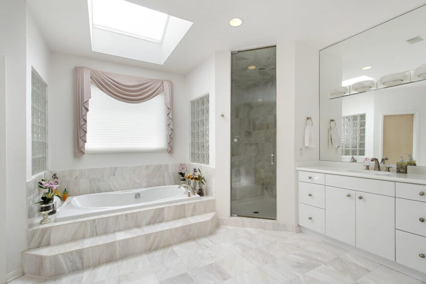 这个光空间的特点是一个彩色玻璃封闭的淋浴间，一个美丽的天窗，和一个相当大的镜子，可以反射光线和空间的错觉。简单的白色橱柜与整个房间的轻石瓷砖相辅相成，而通往放松水疗浴缸的台阶提供了方便。