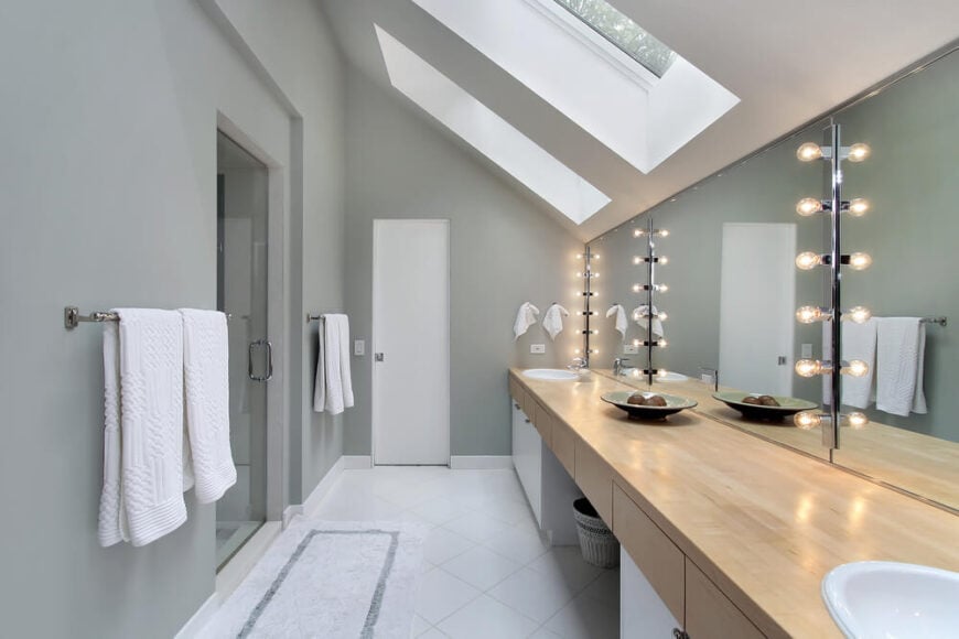 这间现代浴室的哑光灰色墙壁与明亮的白色瓷砖地板形成了美丽的对比。天窗向下照射，带灯的镜子提供额外的氛围。双层梳妆台的天然硬木柜台提供了足够的空间。