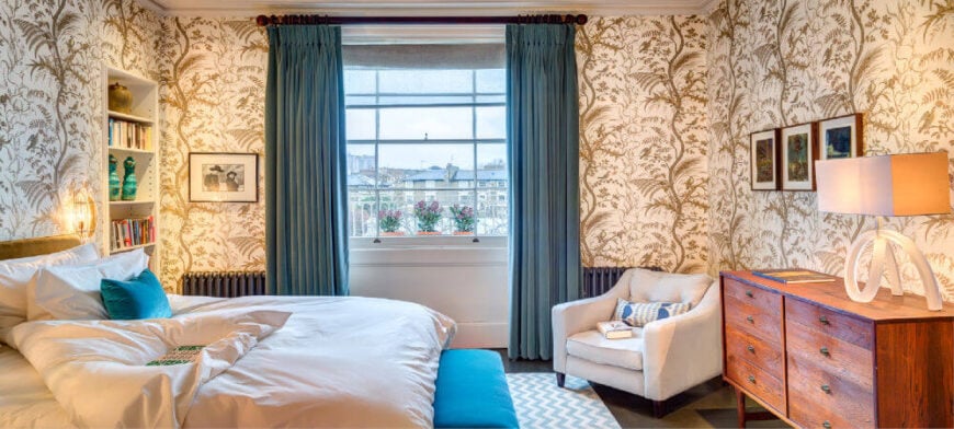 主卧室套房有一个大胆的灰褐色和金色的叶子壁纸，舒适的白色床上用品与蓝绿色的口音。