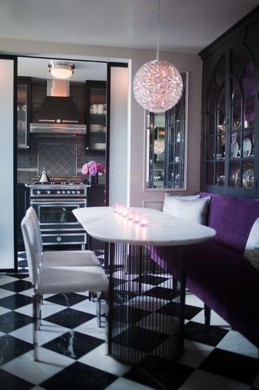 一个令人惊叹的餐厅，一端有一个天鹅绒长凳，对面是内置的瓷器橱柜和两个白色乙烯基餐椅。棋盘格瓷砖地板通过拱门延续到厨房。
