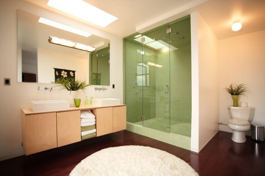 这个吸引人的浴室有一个简单的天窗和照明装置，以最大限度地提高空间内的光线。象牙色的墙壁与深色的硬木形成了美丽的对比，与毛绒圆形地毯形成了进一步的对比。玻璃封闭式淋浴的深橄榄色瓷砖是一个深思熟虑的设计选择，在整个房间中有许多模仿的口音。