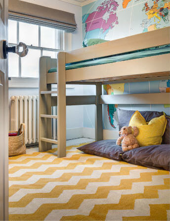 第二个孩子的房间，有黄色的人字形地毯和一张高架床，下面有一个依偎的角落。床后面的墙是一面墙，上面有一幅世界地图。