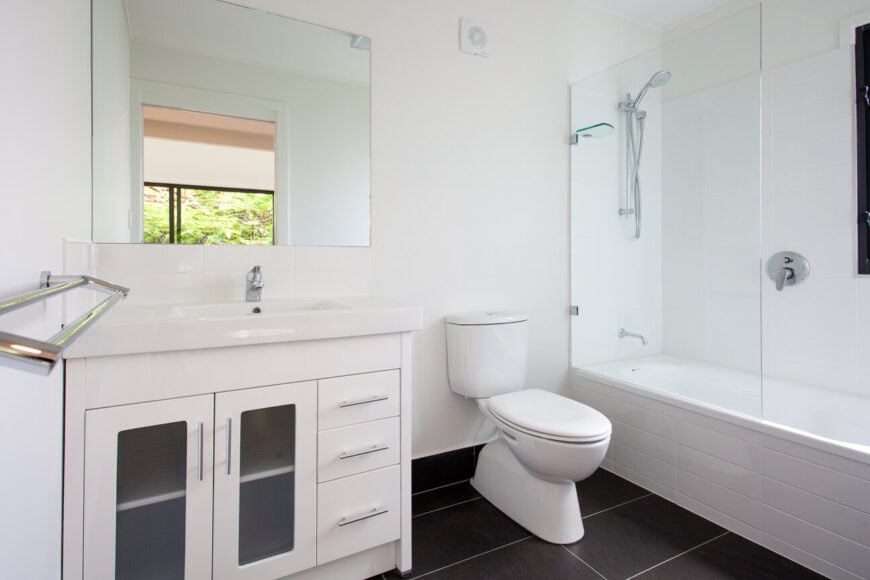 全白色的浴室与深色大面积瓷砖地板形成对比，浴室设有玻璃淋浴间和带烟熏玻璃橱柜的梳妆台。