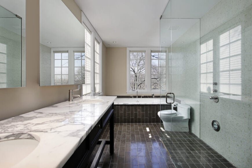 这间时髦的现代浴室采用了褐色的深色砖式瓷砖地板，米色和微瓷砖墙壁下。右边是一间玻璃封闭的大型淋浴间，对面是一间带有大理石台面的台式双梳妆台。
