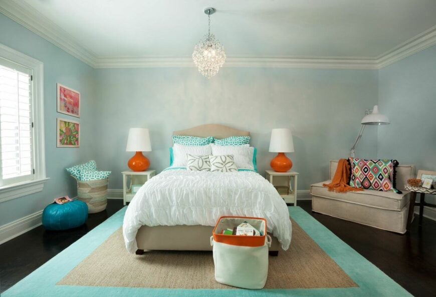 一个别致的现代卧室，柔软的褶皱床上用品和浅蓝色，蓝绿色和橙色的调色板。右边的角落里有一张宽大的长椅，上面有一条柔软的毯子和一个结实的枕头。一盏精致的水晶吊灯悬挂在床的中央。一个可调节的落地灯提供了完美的阅读光线以上的长椅。