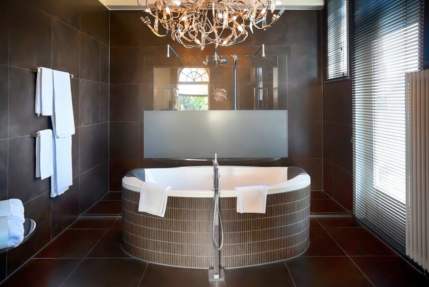 这间方形浴室从地板到地面都包裹着深色铁锈色的大幅瓷砖，中间有一个大浴缸。右边的通高窗为吊灯提供的照明增加了自然光。