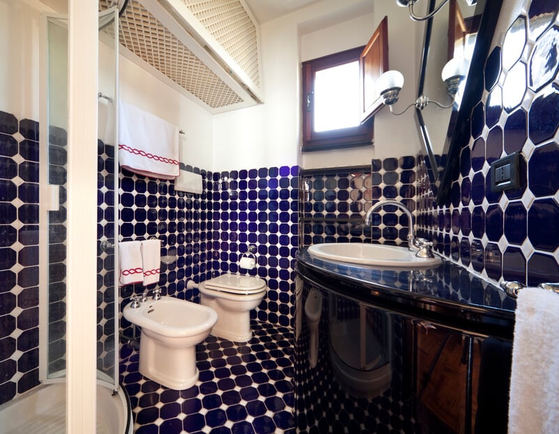 这是另一间独特的蓝色浴室，这一次，蓝色瓷砖从地板一直延伸到墙壁的一半，点缀着白色的点缀。右边是一个弯曲的、光滑的黑色梳妆台，对面是坐浴盆和玻璃封闭的淋浴间。