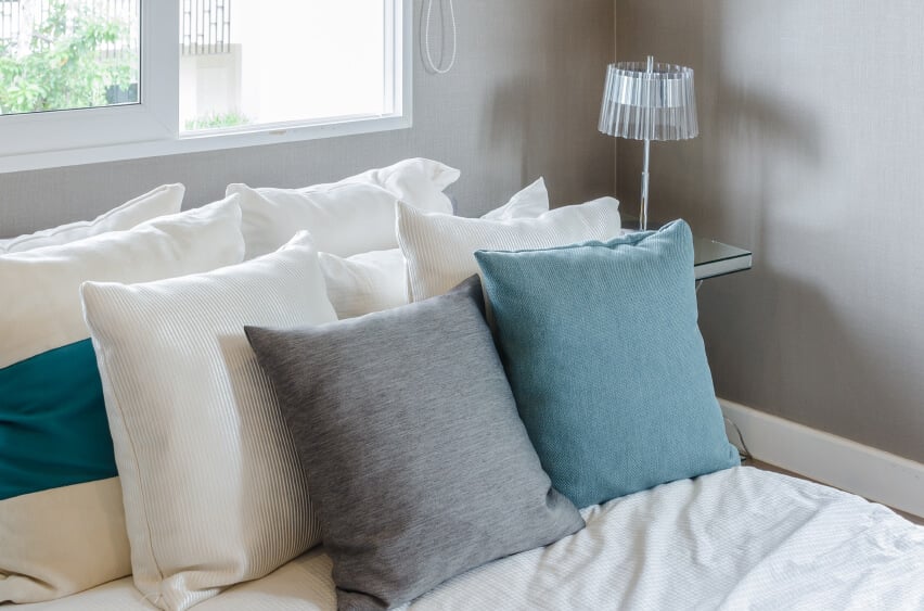 白色的枕头支撑柔软的灰色和蓝色枕头在柔软的床上。灰色的墙壁提供了一个精美的画布，精致的玻璃灯为这个安静的空间带来了额外的兴趣。