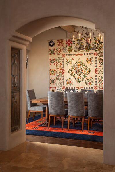 从客厅向餐厅的镜头，可以看到瓷砖与深色硬木地板相遇。定制的餐桌周围摆放着石南灰色软垫的椅子。桌子下面是深海军蓝和橙红色的地毯。后墙上挂着一床巨大的手工棉被。