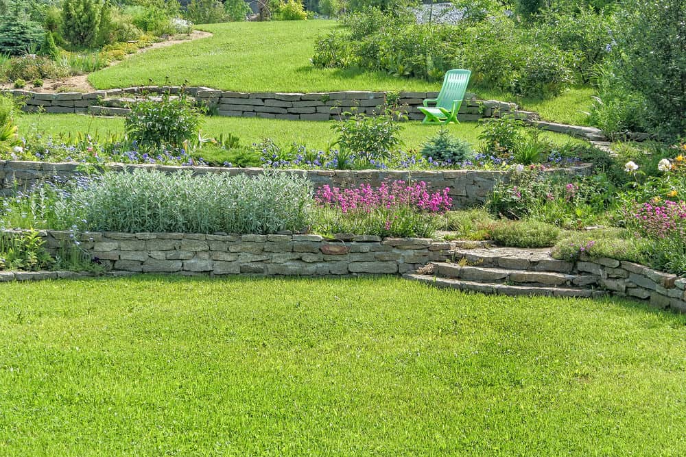 层层叠叠的砖石挡土墙位于郁郁葱葱的斜坡上，可爱的观赏植物强调了这一点，并在第三层配有一把绿色的椅子。