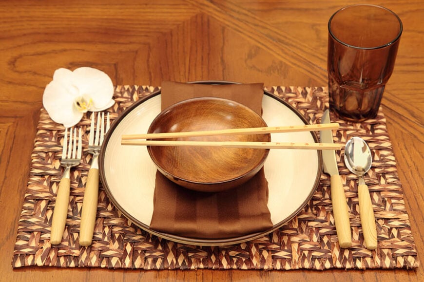 亚洲风格的餐桌摆放在天然纤维餐垫上。左上角摆放着一朵白色兰花，碗上放着一双筷子，旁边放着西方餐具。