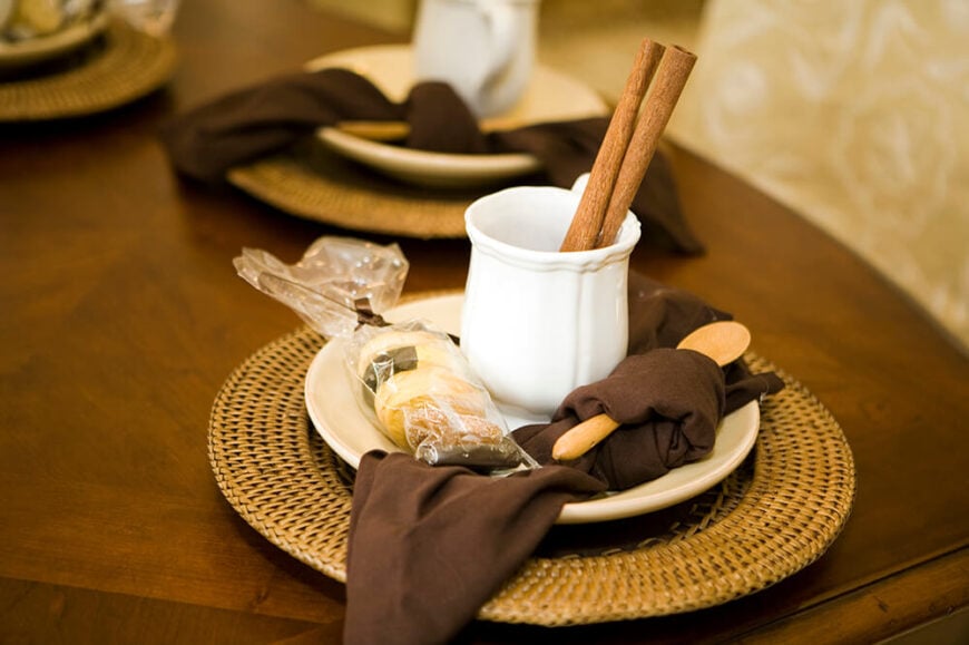 一杯可可的简单桌子摆设。茶托放在柳条托盘里，一个小木勺包在深棕色的餐巾里。旁边有一块蘸酱用的小饼干。