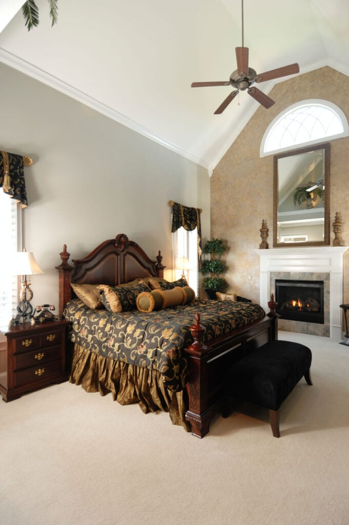更传统的米色卧室，对比鲜明的深色木质家具和丰富质感的床上用品。教堂天花板中央悬挂着一把大扇子。