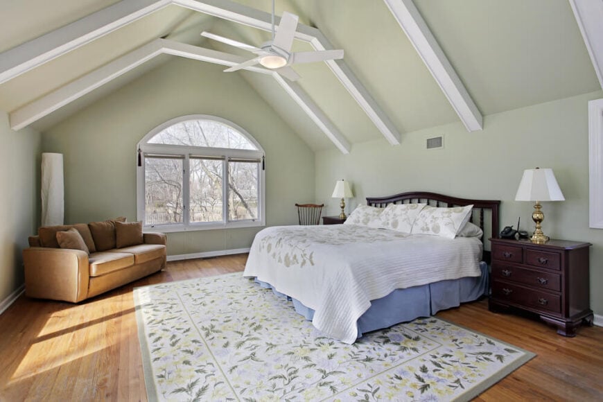 一间简洁迷人的阁楼卧室，裸露的椽子刷成白色，宽敞的窗户旁边有一张单人双人沙发。