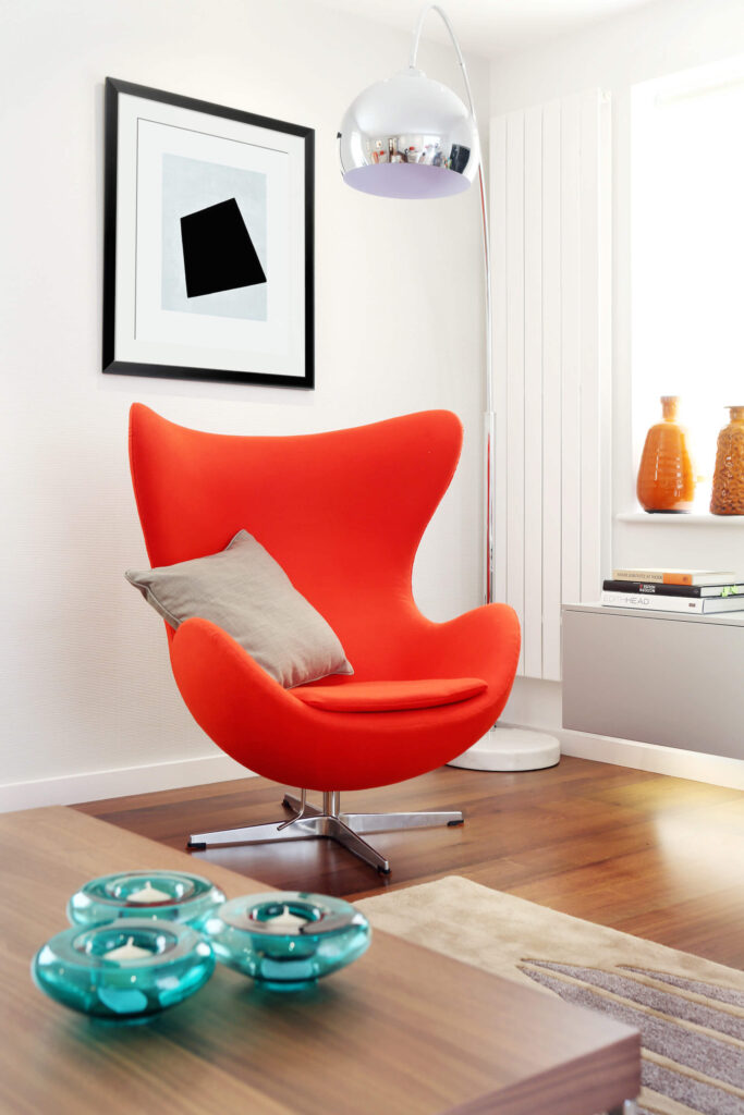 明亮而大胆的橙色椅子，独特的现代鸡蛋形状，增加了巨大的对比和吸引力，特别是与镀铬头顶落地灯相结合。嵌在橱柜里的灯光洒在硬木地板上。
