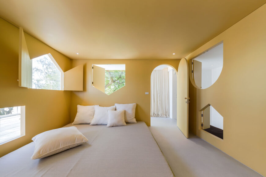 芥末黄色的主卧室展示了各种形状的窗户和一扇拱形门。它有一张灰色的地板床，上面有白色的枕头。