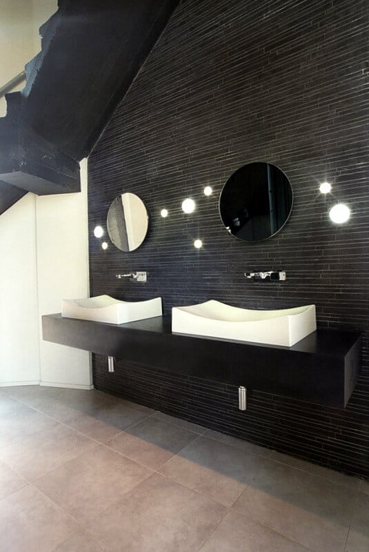 上一层是宽敞的浴室，有一面墙完全贴着长长的黑色瓷砖。明亮的圆球灯点缀在镜子之间的墙上。两个匹配的容器水槽在极简主义的黑色梳妆台上呈现出感官上的曲线。
