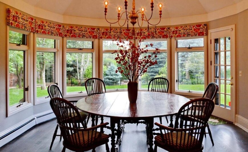 在厨房旁边，我们有一个更亲密的家庭用餐空间，一个由通高窗户环绕的圆形房间，可以看到广阔的花园景观。一盏老式的枝形吊灯下放着一张传统的大木制圆桌。