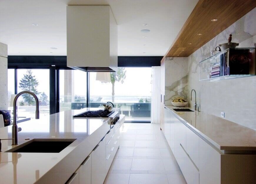 这个光滑的现代厨房通过环绕玻璃，浅色瓷砖地板，墙壁和台面看起来明亮。漫长的极简主义岛设有一个完整的二级水槽以及一个大型不锈钢燃气灶台。