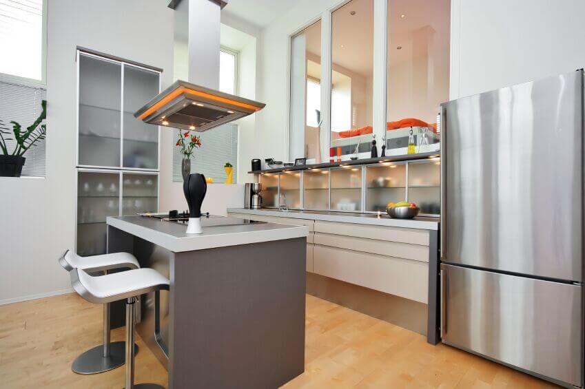 这是一个特别明亮的厨房，充斥着各种各样的纹理，从浅色硬木地板到烟熏玻璃橱柜。大型极简主义岛台的特点是灰色台面，表面平坦，头顶上包裹着橙色条纹的独特抽油烟机。