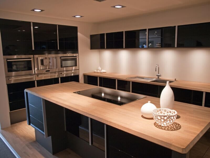 这个光滑，高对比度的现代厨房设有光滑的黑色橱柜，并与浅色天然木质台面和地板并列。大岛台拥有一个玻璃台面，与橱柜的黑色色调相匹配。