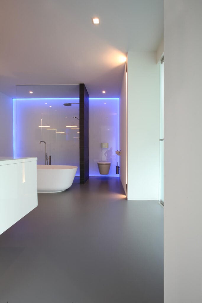 锃亮的白色浴室里有一间大型玻璃淋浴间和时尚的基座浴缸，带有未来主义风格。一盏隐藏的霓虹灯环绕着浴室远壁的四周，为浴室增添色彩。