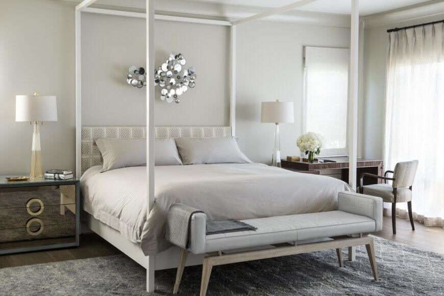 这张四柱床的薄白色柱子吸引了人们对大床的注意，并为浅灰色的墙壁增添了视觉趣味和光线对比。镜子床头柜和一张小桌子为房间增添了更多的质感和光线。