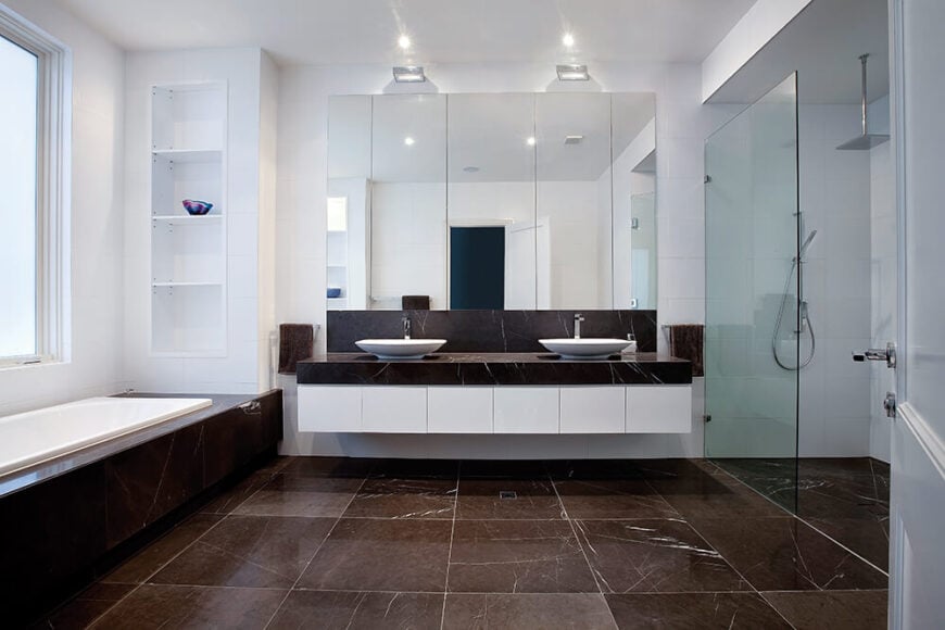这间主浴室铺着一大片深色大理石地板，右边是一间玻璃封闭式步入式淋浴间，左边是窗户下方的大浴缸。漂浮的双梳妆台以白色橱柜和大理石表面为特色，支撑着一对白色容器水槽。
