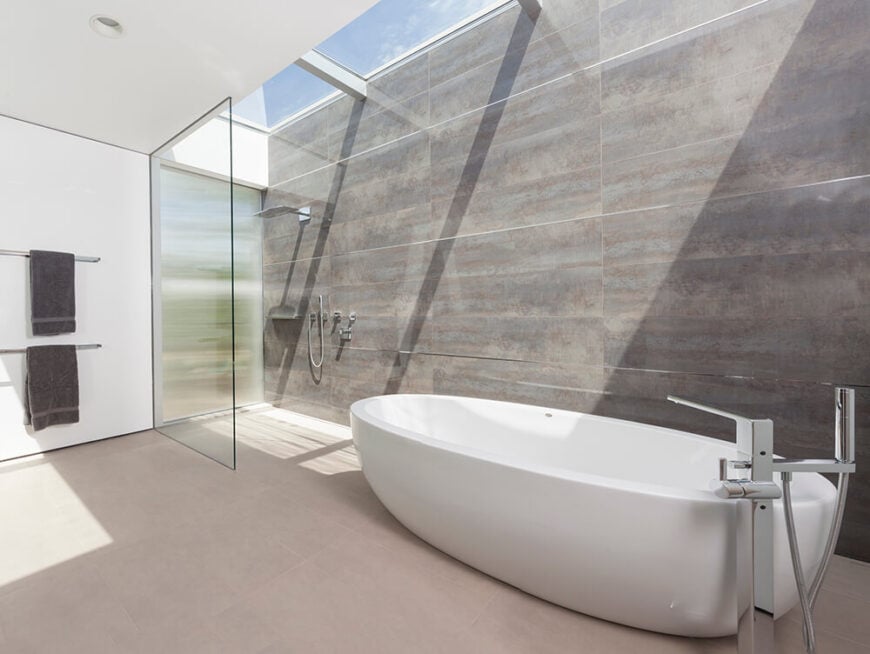 主浴室是一个巨大的极简主义空间，充满了混凝土、玻璃和白墙。一组房间长度的天窗确保了白天的自然光线，在步入式淋浴间和大型白色底座浴缸上发光。