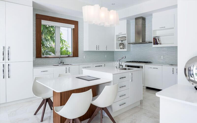 一间白色的现代厨房，后挡板是浅灰色的玻璃马赛克瓷砖，还有一个小岛，用作厨房内的用餐区、额外的台面和存储空间。