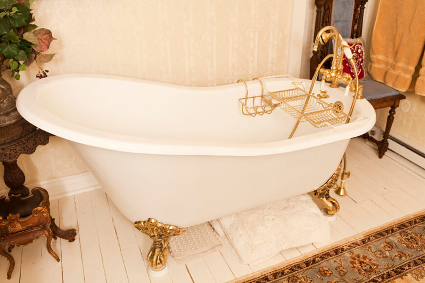 金色的爪形浴缸坐落在白色瓷砖地板上，大面积的地毯似乎覆盖了大部分地板空间。浴缸旁的一张小桌子上摆放着装饰性植物，与浴室的墙纸相得益彰。