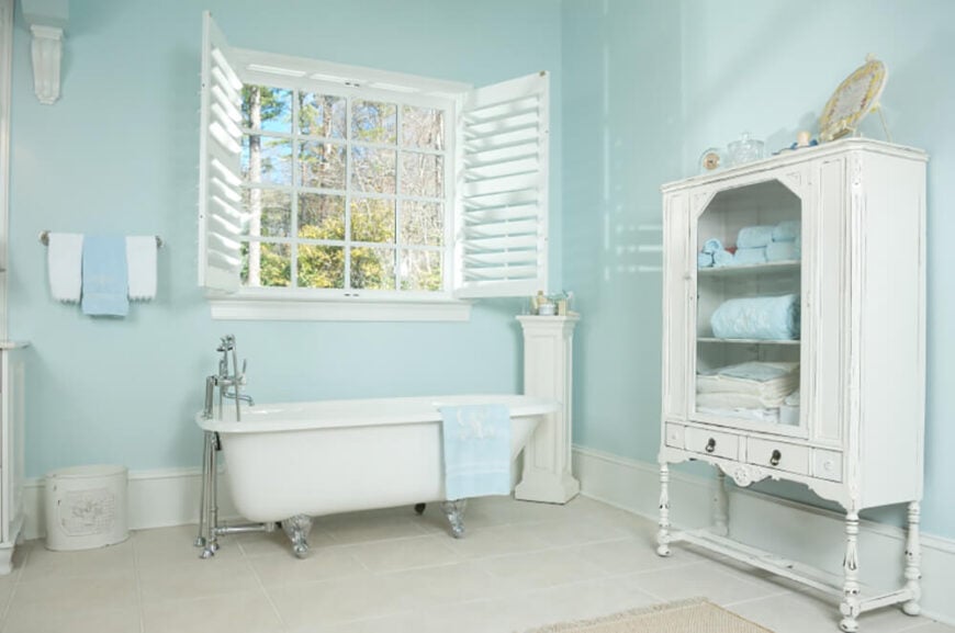 双百叶窗打开，让自然光透过这扇窗户照进来。窗户正下方有一个爪形浴缸。浅蓝色的墙壁和特色毛巾与白色的地板和公用设施相结合，使这个空间明亮而温馨。
