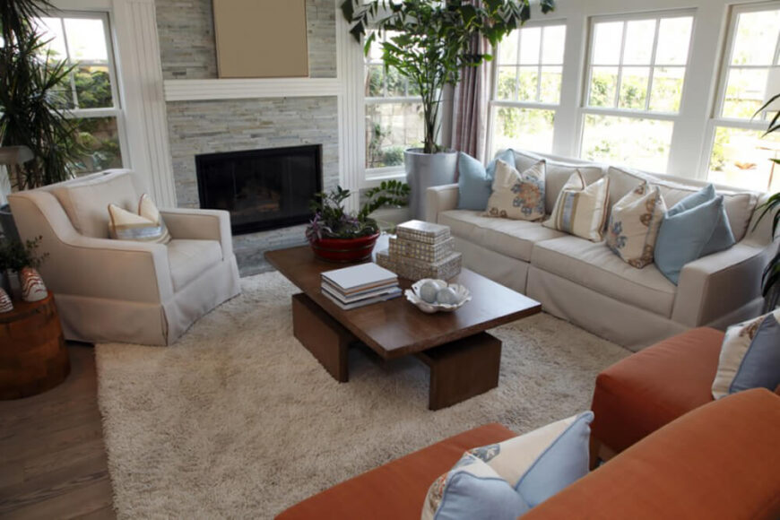 室内植物为这个宁静的房间增添了宁静的气氛。现代家具搭配层次化的石材壁炉和柔和的木地板，巩固了空间的设计。枕头和两个边椅的焦橙色色调在中性色调中创造了兴趣。
