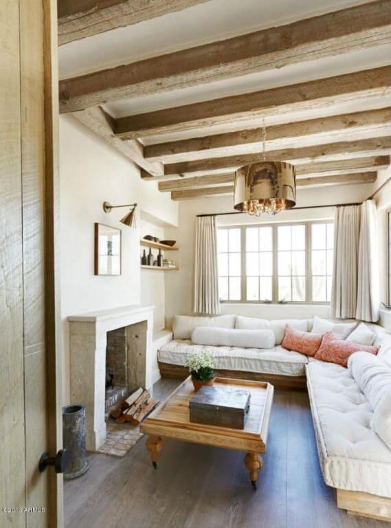 这间舒适的客厅融合了现代色调和乡村魅力，上面有裸露的天然木梁，下面有丰富的硬木地板。白色软垫组合家具包裹着两面墙，拥抱着面对壁炉的箭尺咖啡桌。