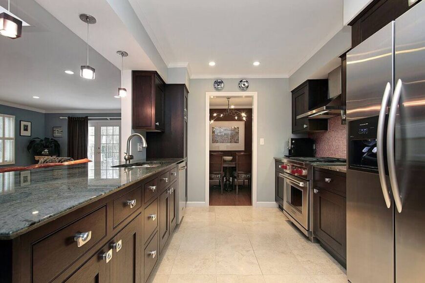 这个现代厨房的布局完全是长方形的，以绿灰色的花岗岩台面、深色木橱柜和与不锈钢高端电器相匹配的银色现代固定装置为特色。厨房俯瞰着深蓝色的客厅区，是娱乐的好设计。