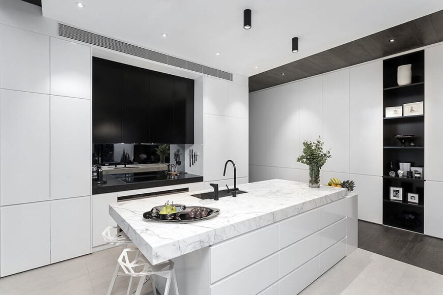厨房本身是一个白色的大岛台，带有厚厚的白色大理石台面和嵌入式水槽。黑色系列两侧是光滑的白色橱柜，右边是一个细长的展示架。