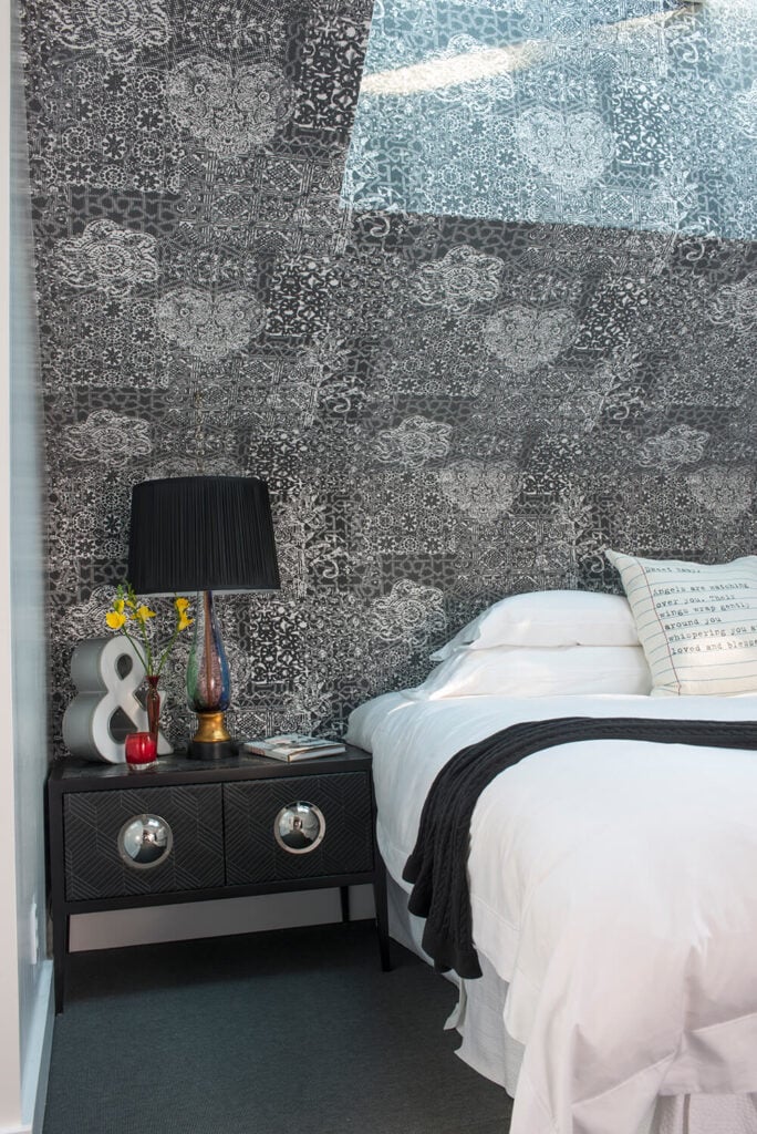 床头柜有独特的镀铬硬件和内部存储，支持艺术作品，如彩色玻璃灯。细致的墙面纹理与朴素的白色床上用品和灰色地毯形成鲜明对比。