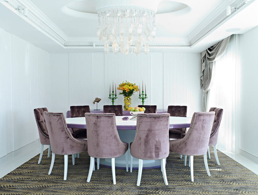 餐厅的墙壁和天花板上有微妙的纹理，一个独特的枝形吊灯悬挂在巨大的紫色边缘桌子上。一套钉头装饰的Parson椅子将桌子包裹在印花地毯上，形成高对比度。