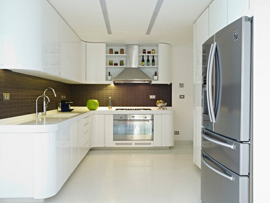 厨房里到处都是优美的曲线和光滑的表面，白色的橱柜和台面铺着白色的瓷砖地板。微妙的深色地铁瓷砖后挡板和不锈钢器具提供了对比和纹理。