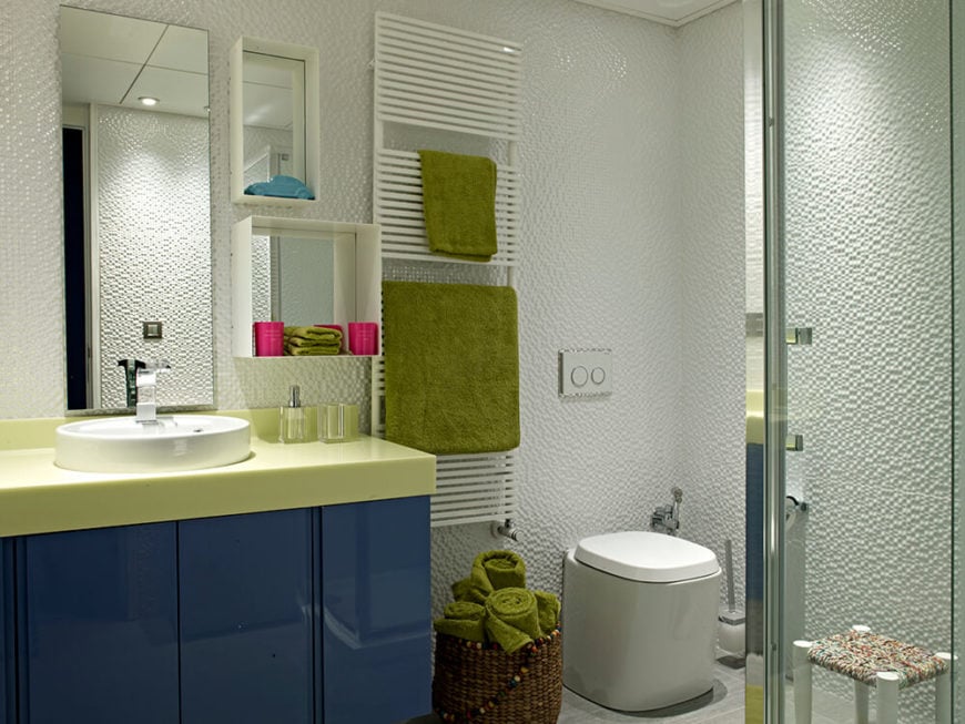 这是第二间浴室，风格与第一间完全不同。深蓝色和黄色的梳妆台位于一系列包含架子的镜子下方，而高度纹理的墙壁使白色空间更加细致。